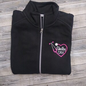 May include: Black fleece zip-up jacket with a pink embroidered stethoscope and heart with the text "Shelby CMA".