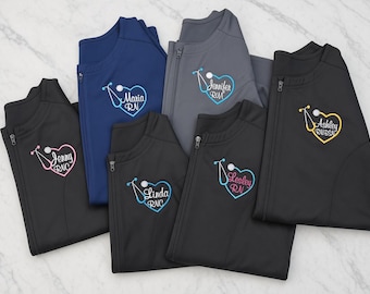 Puede incluir: Seis chaquetas con cremallera en negro, azul marino y gris con logotipos bordados en el pecho. Cada logotipo muestra un estetoscopio en forma de corazón con un nombre y título médico, incluyendo Jenny RNC, Maria RN, Jennifer RN, Linda RNC, Lesley RN y Ashley RN-BSN.