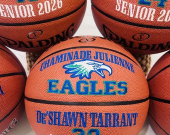 May include: Five orange Spalding basketballs featuring black seams and custom embroidery. The centre ball displays the Chaminade Julienne Eagles logo, the name De'Shawn Tarrant, the number 32, and Senior 2026. Other balls show names, numbers, and graduation years.