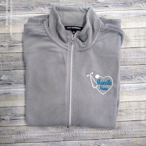 May include: Gray fleece zip-up jacket with a white zipper and a blue embroidered stethoscope and heart with the text "Marcella Nurse".