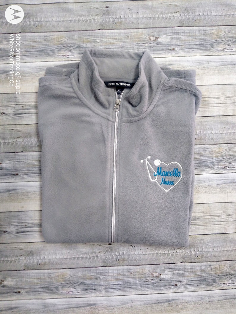 May include: Gray fleece zip-up jacket with a white zipper and a blue embroidered stethoscope and heart with the text "Marcella Nurse".