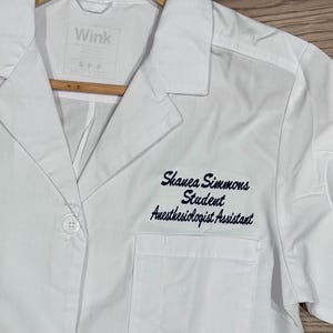 May include: White lab coat with a blue embroidered name tag that reads "Shanea Simmons Student Anesthesiologist Assistant". The coat has a pocket on the left side and two buttons on the front.