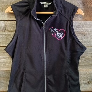 May include: Black fleece vest with a zipper closure and a pink embroidered heart with the text "Grace RN" and a stethoscope.