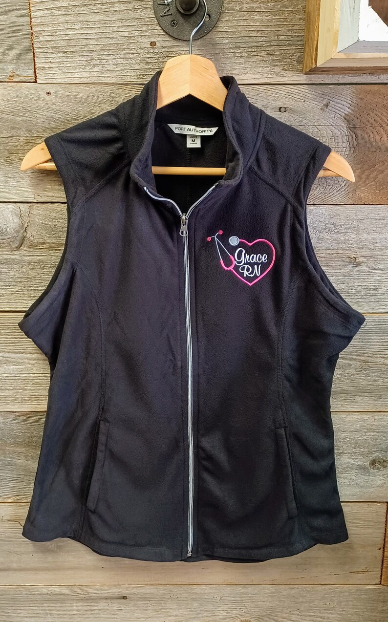 May include: Black fleece vest with a zipper closure and a pink embroidered heart with the text "Grace RN" and a stethoscope.