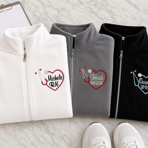May include: Three fleece zip-up jackets in white, gray, and black, each embroidered with a heart-shaped stethoscope design and personalized text. The white jacket reads Michele RN, the gray reads Heidi RN, BSN, and the black reads Quanisha LPN.