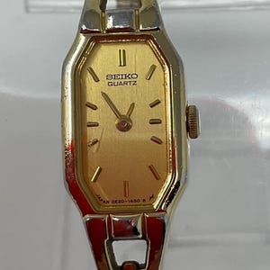 May include: A gold-toned Seiko Quartz wristwatch with a rectangular face. The watch features a gold dial with black markers and hands. The band is a gold and silver tone. The watch face has the text "SEIKO QUARTZ" and "JAPAN 2E20-1450 R".