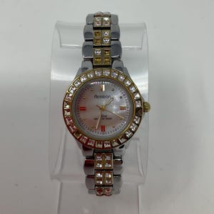 Puede incluir: Un reloj Armitron plateado y dorado con esfera de nácar. El reloj presenta un bisel y una correa adornados con cristales transparentes. El reloj tiene una esfera redonda con agujas y marcadores dorados.