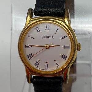 May include: A gold-plated Seiko wristwatch with a white face and black Roman numerals. The watch has a black leather strap and gold hands. The watch face displays the brand name "SEIKO".