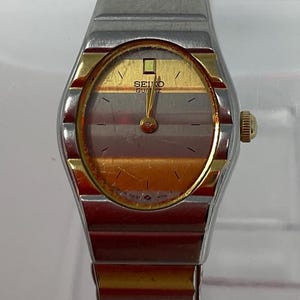 May include: A vintage Seiko Quartz watch with a gold and silver-toned metal band and case. The watch face is oval-shaped with a gold and brown striped design. The watch has a gold crown and hands.