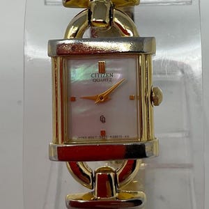 May include: A gold-toned Citizen Quartz watch with a rectangular face and a mother-of-pearl dial. The watch features gold hands and hour markers, and a gold-toned band. The watch face displays the Citizen Quartz logo.