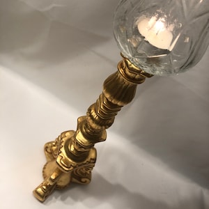 Könnte beinhalten: Goldener Kerzenhalter mit einem klaren Glas-Windlicht. Der Halter hat ein detailliertes, verziertes Design und eine breite Basis.