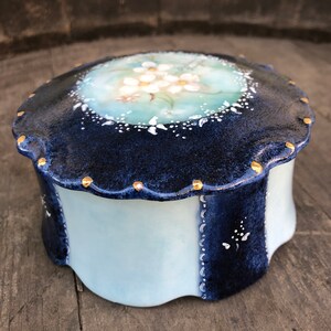 May include: A small, round, blue and white ceramic trinket box with a scalloped edge and gold accents. The lid features a floral design with white flowers on a light blue background.