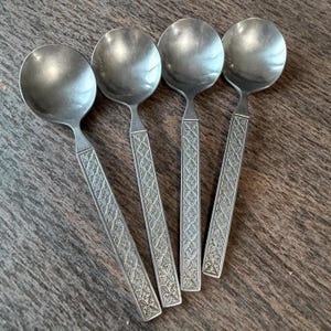 May include: Four vintage silver spoons with round bowls and patterned handles. The spoons are arranged on a wooden surface, showcasing their reflective surfaces. The handles feature a detailed, textured design.