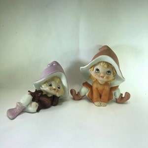 May include: Two ceramic figurines of elves with pointed hats. The elf on the left is laying down with a purple hat and brown and white clothing. The elf on the right is sitting up with a brown hat and orange clothing.