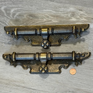 May include: Two antique bronze-toned cabinet pulls with ornate detailing. Each pull features a central decorative element and a curved, stylized base. A penny is included for size reference. The hardware has a vintage aesthetic.