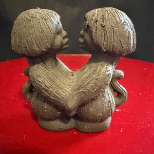 May include: A dark gray clay sculpture depicting two figures with bobbed hairstyles facing each other. The figures have textured surfaces and are joined at the torso, with their arms wrapped around each other in a connected, symmetrical pose.