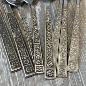 Op de afbeelding: Close-up van een set vintage zilveren besteklepels. De handvatten zijn zilverkleurig en hebben een bloemreliëf. De lepels zijn gerangschikt op een lichtgrijze houten ondergrond.