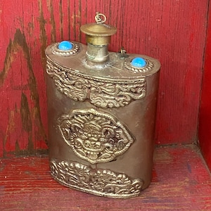 Può includere: Una fiaschetta in metallo argentato e dorato con intricati disegni in rilievo e due pietre turchesi blu sul coperchio. La fiaschetta ha un coperchio incernierato con un piccolo anello in cima.