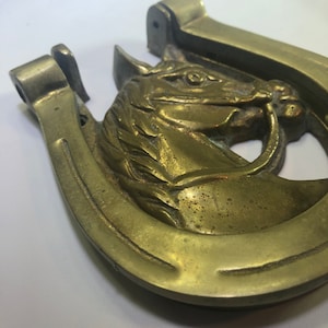 May include: A brass horseshoe door knocker featuring a horse's head design. The horseshoe is a traditional symbol of good luck.