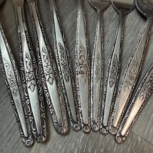 Puede incluir: Una colección de cubiertos antiguos con un diseño floral de rosas. Los utensilios de plata incluyen tenedores y cucharas, con intrincados patrones de rosas y hojas en relieve a lo largo de los mangos. La cubertería está dispuesta sobre una superficie de madera.