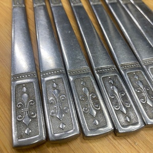 May include: Close-up of eight silver flatware pieces with ornate designs. Each piece has a decorative pattern of a fleur-de-lis and a diamond shape.