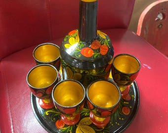 May include: A black wooden tray with a black and gold painted bottle and six small cups. The bottle and cups are decorated with red and yellow flowers and berries.