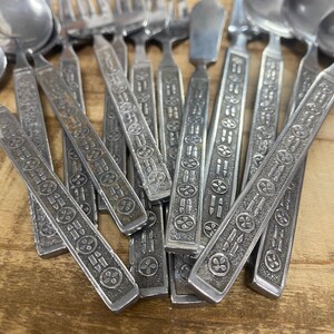 May include: A collection of vintage silverware with decorative handles. The stainless steel utensils include forks, spoons, and butter knives. The handles feature an embossed pattern of circles and lines. The silverware is arranged on a wooden surface.