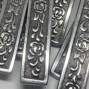 Op de afbeelding: Close-up van verschillende zilveren rechthoekige metalen stukken met een bloemenrozenontwerp. De rozen en bladeren zijn verhoogd tegen een getextureerde achtergrond. De stukken hebben een vintage of antieke uitstraling.