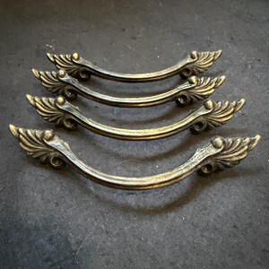 May include: Four curved metal drawer pulls with an antique brass finish. Each handle features a central arch and decorative wing-shaped ends with detailed ridges. The hardware has a weathered, dark patina, showcasing a consistent design across all pieces.