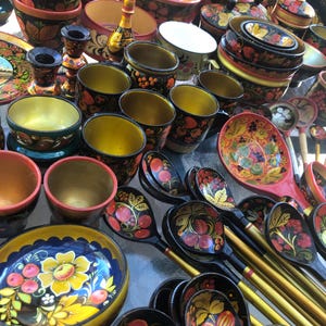 Puede incluir: Una colección de cuencos, tazas y cucharas de madera pintadas a mano con intrincados diseños florales en rojo, amarillo y verde. Los cuencos y las tazas están anidados, mientras que las cucharas están dispuestas en forma de abanico.