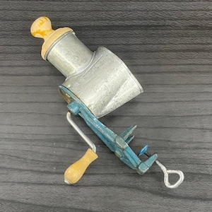 Puede incluir: Un molinillo de alimentos vintage de metal con una manivela de madera y una base de metal azul. El molinillo tiene un cuenco de metal plateado con una perilla de madera en la parte superior.
