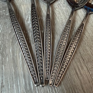 May include: A close-up shot of a collection of silver-coloured cutlery. The handles are decorated with a detailed leaf pattern. The spoons and serving utensils are arranged on a wooden surface, highlighting the intricate patterns and textures.