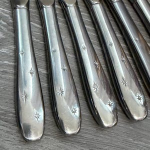 May include: A set of silver-toned metal flatware handles arranged in a row. Each handle features a smooth, polished surface decorated with small, engraved starburst patterns scattered along the length of the handle, creating a consistent design.