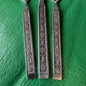 May include: Close-up of three silver-colored utensils with decorative floral and geometric patterns on the handles. The handles are rectangular and feature alternating floral and geometric designs. The utensils are set against a green background.