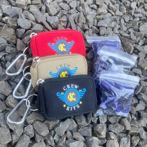 May include: Three zippered pouches with a keychain, each with a different color: red, beige, and black. The pouches have a logo with the text "CREW" and a stylized "KC" with wings. The pouches are on a background of small gray rocks.
