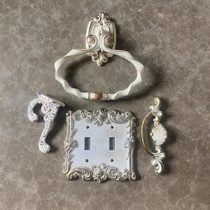 May include: Set of five ornate white and gold metal hardware pieces, including a towel ring, a hook, a double light switch cover, and two drawer pulls.
