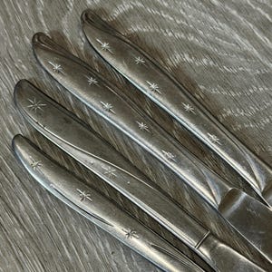 May include: Close-up of several silver knives with starburst designs. The knives are arranged diagonally, showcasing their polished surfaces and decorative details. The handles feature a series of star-shaped engravings.