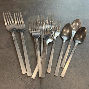 Könnte beinhalten: Eine Sammlung von Vintage-Besteck, darunter Gabeln und Löffel, auf einer grauen Oberfläche angeordnet. Das Besteck hat ein Blumendesign auf den Griffen. Die Gabeln haben vier Zinken und die Löffel haben ovale Schalen.