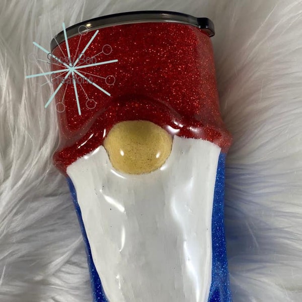 May include: A red, white, and blue glitter tumbler with a gnome face design. The gnome has a white beard, a red hat, and a yellow nose.