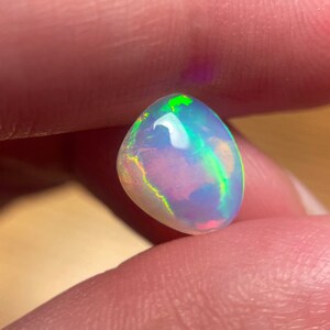 May include: A white opal gemstone with green, blue, and pink iridescent flashes. The opal is a teardrop shape and is held in a hand.