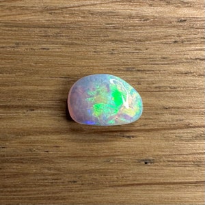 May include: A white opal gemstone with green and blue iridescent flashes. The opal is a freeform shape and has a smooth, polished surface.