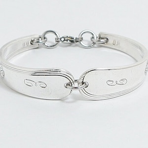 May include: A silver bracelet crafted from repurposed silverware. The bracelet features a series of spoon-shaped links connected by small rings. The design includes engraved details and a secure clasp. The bracelet is approximately 7 inches in length.