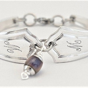 May include: Silver bracelet made from two spoons, joined together with a chain. The spoons have the initials "MM" engraved on them. A purple and gray bead hangs from the bracelet.