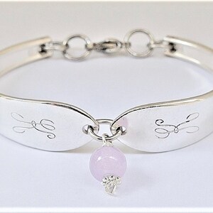 May include: A silver bracelet made from a repurposed spoon with a delicate charm featuring a light pink gemstone. The bracelet has a clasp and is engraved with the initials "S & S".