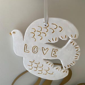 May include: White dove-shaped ornament with gold detailing and the word "LOVE". The wings and tail feathers have gold lines. A white ribbon is attached for hanging. The ornament is likely ceramic or plaster.