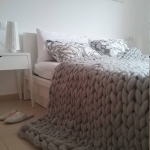 May include: A gray, chunky knit blanket draped over a white bed. The blanket is made of thick, soft yarn and has a textured appearance. Two decorative pillows with a gray and white floral pattern are on the bed. A white nightstand and a decorative lamp are visible.