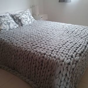 May include: A large, chunky-knit gray blanket covers a bed, paired with two decorative pillows featuring a white and gray floral pattern. The bed is made up with white sheets. A pair of gray slippers rests on the floor.