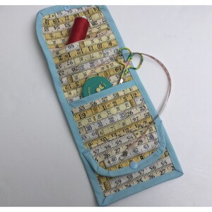 May include: A light blue fabric sewing kit with a zipper closure. The kit is open and contains a red spool of thread, a small green tape measure, and a pair of silver scissors. The kit is lined with yellow fabric with a pattern of measuring tape.