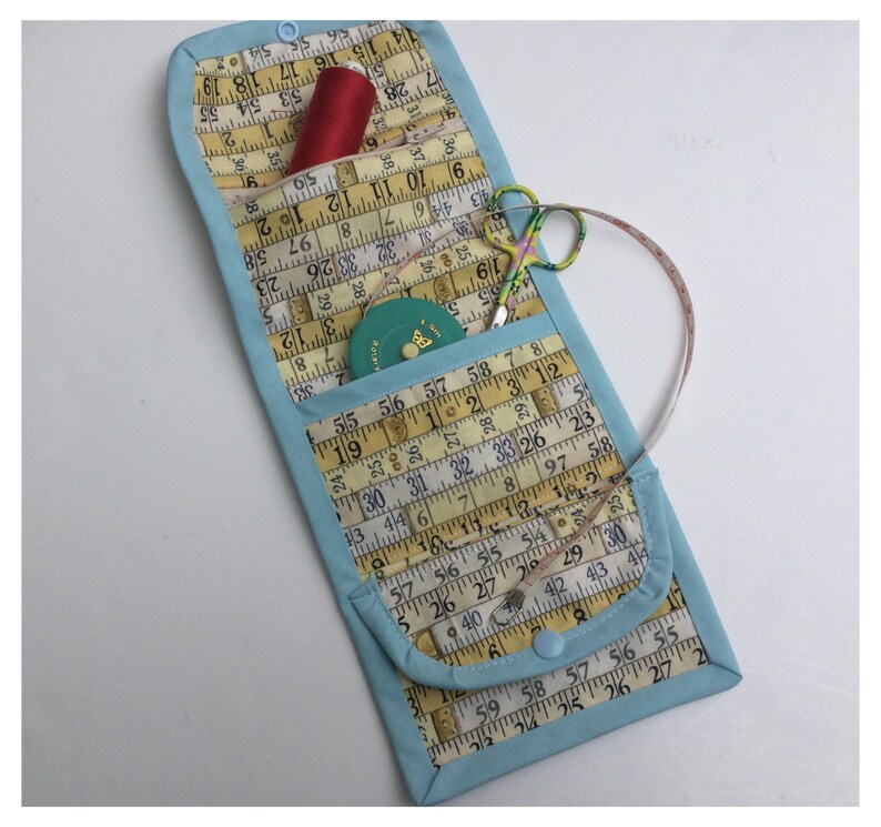 May include: A light blue fabric sewing kit with a zipper closure. The kit is open and contains a red spool of thread, a small green tape measure, and a pair of silver scissors. The kit is lined with yellow fabric with a pattern of measuring tape.