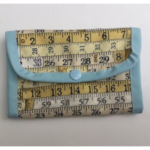 May include: A light blue fabric wallet with a snap closure. The wallet is made from fabric with a pattern of yellow measuring tapes with black numbers. The numbers on the tape measure include 2, 3, 4, 5, 6, 7, 8, 26, 27, 28, 29, 30, 31, 32, 33, 55, 56, 57, 58, 59, 12, 13, 14, 15, 16, 17, 23, 24, 25, 26, 27.